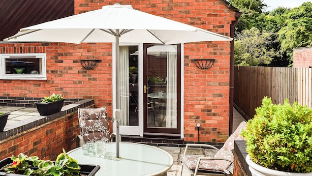 Une terrasse avec une table et des chaises en verre, un parasol blanc et une maison en briques dotée d’une fenêtre et d’une porte.