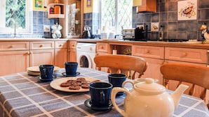 Une cuisine avec des meubles en bois, une table dressée avec des tasses bleues et une théière, et une fenêtre donnant sur un paysage verdoyant.