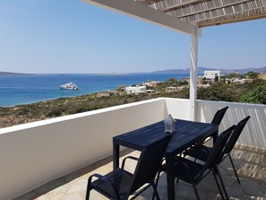 Ein Balkon mit Tisch und Stühlen, der auf eine Küstenlandschaft mit einem Segelboot in der Ferne blickt.