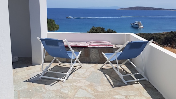 En terrass med två blå solstolar, ett bord med en randig kudde och utsikt över havet med båtar.