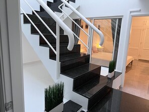 Escadaria moderna com degraus pretos e corrimãos brancos, que dá acesso a um quarto com uma janela grande e um abajur.