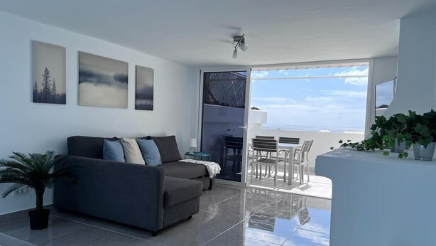 Uma sala de estar moderna com um sofá, um espaço de jantar com cadeiras e vista para o mar.