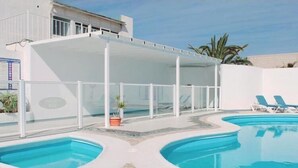 Uma piscina com duas áreas distintas, uma área de estar coberta com cadeiras e um edifício ao fundo.