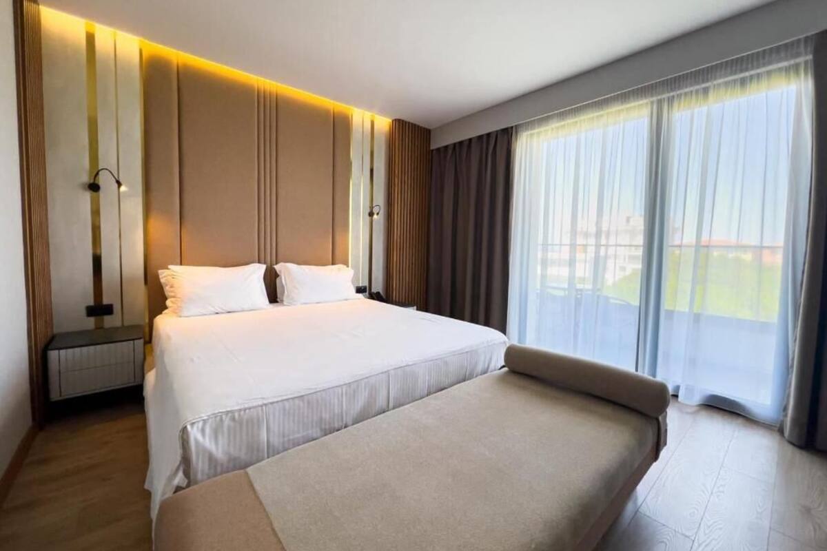A modern hotel room with a large bed, a bedside table, a lamp, and a large window with curtains.