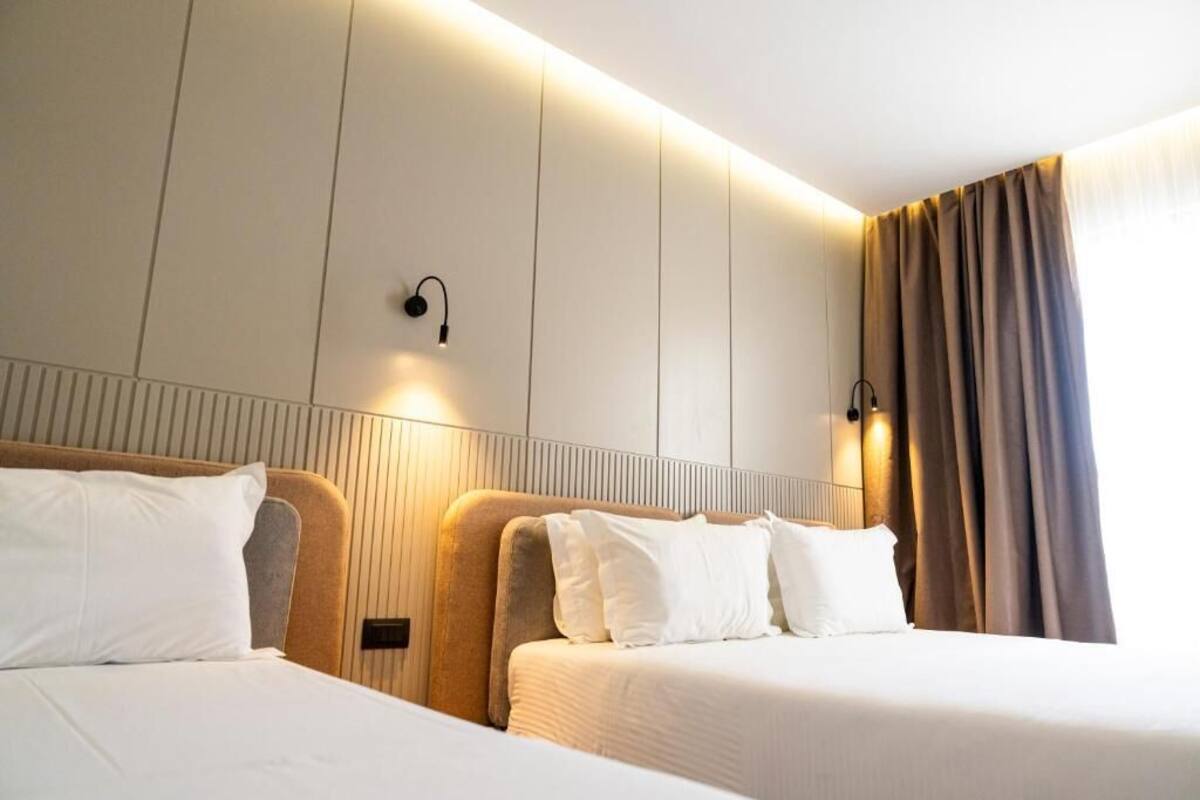 A hotel room with two beds, a headboard with integrated lighting, a wall-mounted light fixture, and a window with curtains.