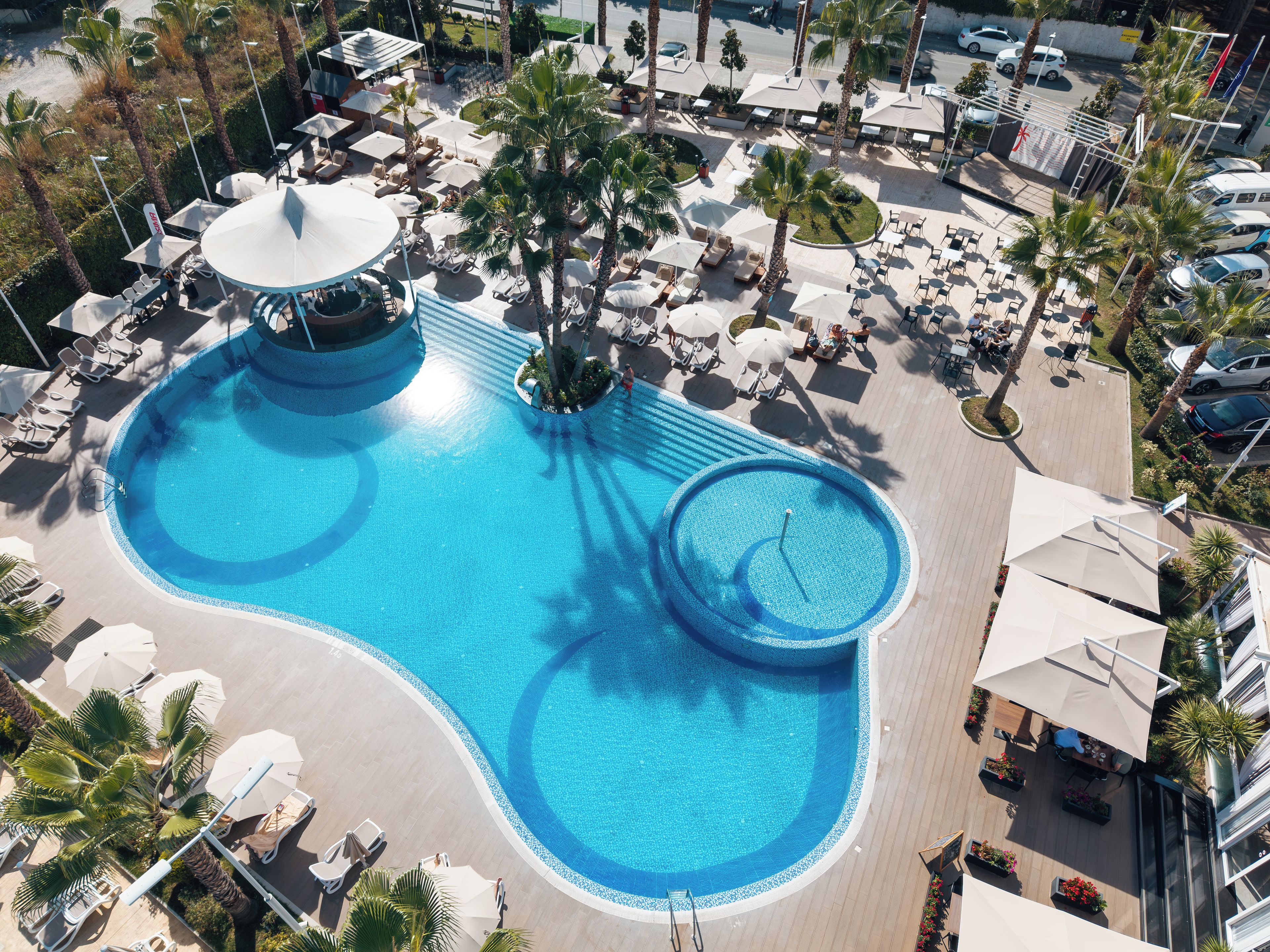 Vista aérea de un complejo turístico con una gran piscina central, varias piscinas más pequeñas y numerosas tumbonas y sombrillas.