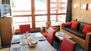 Un coin repas avec une table dressée pour deux personnes, des chaises, un canapé avec des coussins rouges et un téléviseur posé sur un meuble en bois.