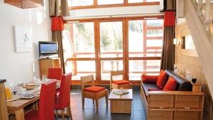 A modern hotel room with a wooden dining table, red chairs, a sofa with orange cushions, and large windows offering a view of the outdoors. - travelski home premium - Résidence Le Roc Belle Face 4* - 3 pièces alcôve et/ou duplex 8 personnes (Les Arcs)