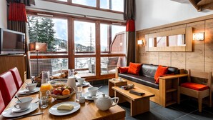 A cozy room with a dining table set for breakfast, a sofa with cushions, and a TV on a stand. - travelski home premium - Résidence Le Roc Belle Face 4* - 3 pièces alcôve et/ou duplex 8 personnes (Les Arcs)
