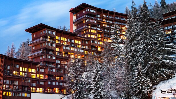 A multi-story hotel with balconies and warm lighting, surrounded by snow-covered trees. - travelski home premium - Résidence Le Roc Belle Face 4* - 3 pièces alcôve et/ou duplex 8 personnes (Les Arcs)