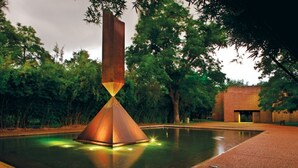 Une sculpture moderne se reflète dans un bassin, entourée d’une zone pavée et avec des arbres en arrière-plan.