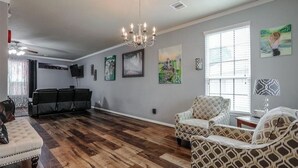 Un salon spacieux doté d’un parquet, d’un lustre et d’une grande fenêtre avec des stores.
