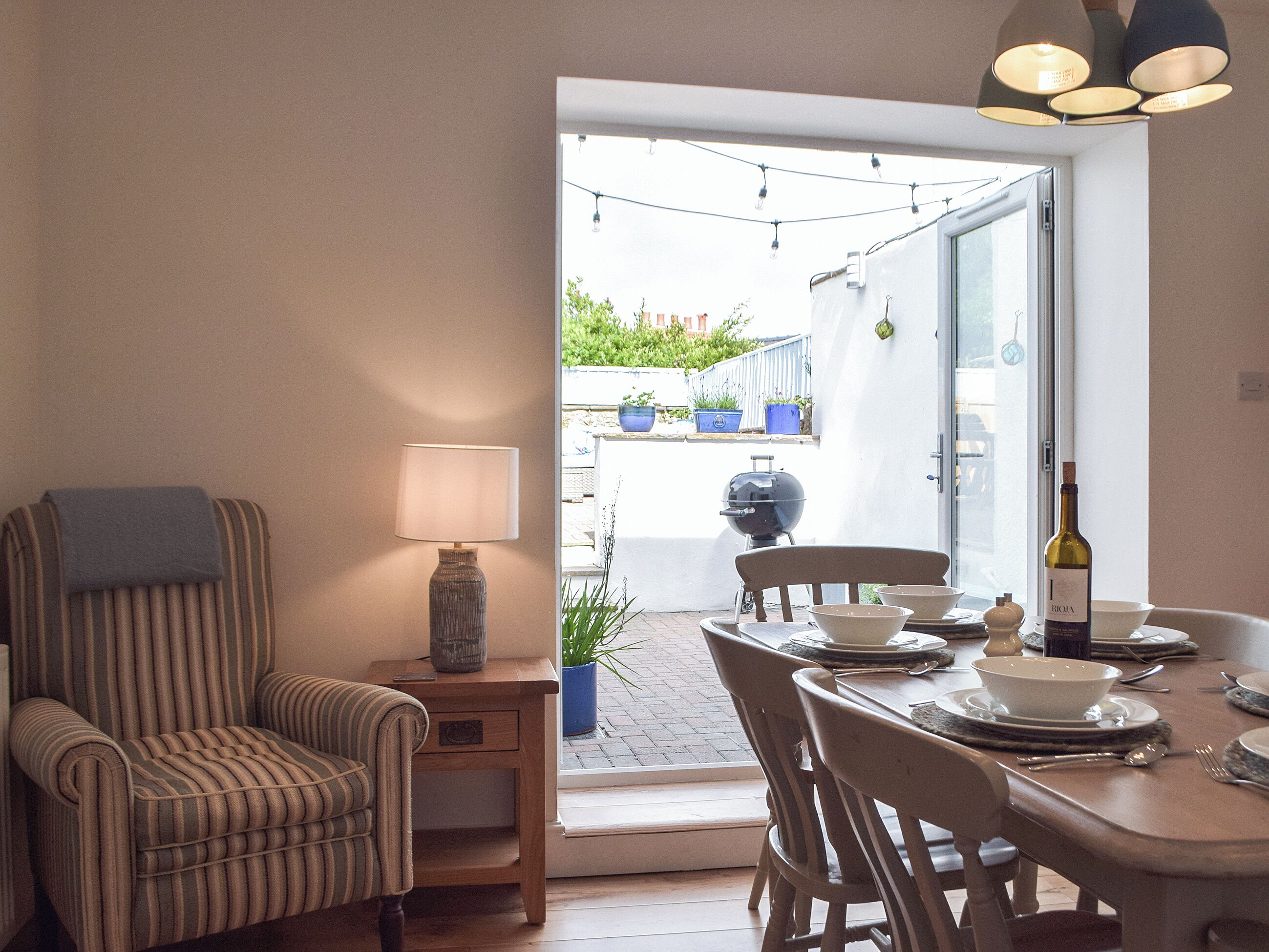 A dining area with a table set for four, a striped armchair, a small side table with a lamp, and a bottle of wine.