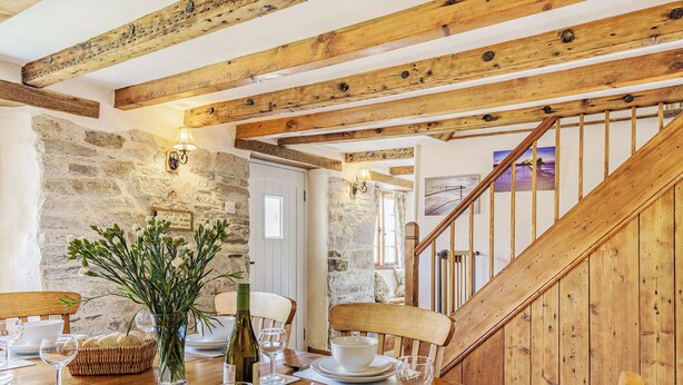 A wooden dining table set with plates, glasses, and a bottle, in a room with exposed wooden beams and stone walls. - A charming, detached farmhouse with plenty of character. (St Columb Major)