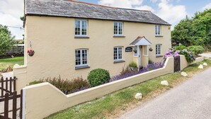 A two-story building with a gabled roof, a small porch with a sign, and a flower bed with purple flowers. - A charming, detached farmhouse with plenty of character. (St Columb Major)