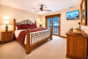 A bedroom with a wooden bed, two bedside tables with lamps, a ceiling fan, a large window with a view, and a dresser with decorative items.