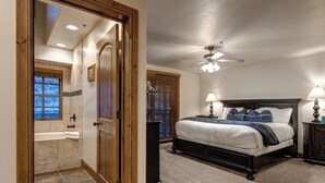 A bedroom with a bed, a ceiling fan, a nightstand, a lamp, a window with blinds, and a door leading to a bathroom.