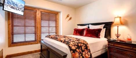 A bedroom with a bed, a bedside table with a lamp, a dresser, and a wall-mounted TV.