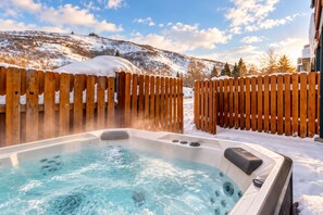 Een hottub omringd door houten hekken, met sneeuw op de grond en een bergachtige achtergrond.