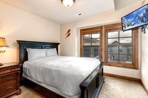 Un dormitorio con una cama, una mesita de noche, una lámpara, una ventana con persianas y un televisor montado en la pared.