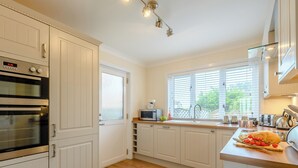 A modern kitchen with white cabinetry, a built-in oven, a window with blinds, and a wooden countertop with a loaf of bread and tomatoes. - 4 bedroom accommodation in Beadnell (Beadnell)