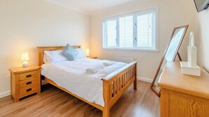 A bedroom with a wooden bed, bedside tables, a nightstand, a window with blinds, and a mirror on a dresser. - 4 bedroom accommodation in Beadnell (Beadnell)