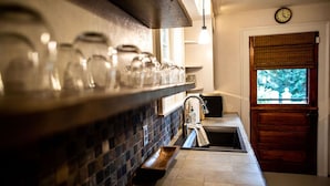 A kitchen with a brick backsplash, a wooden shelf with glassware, a sink, and a window with a view of greenery.