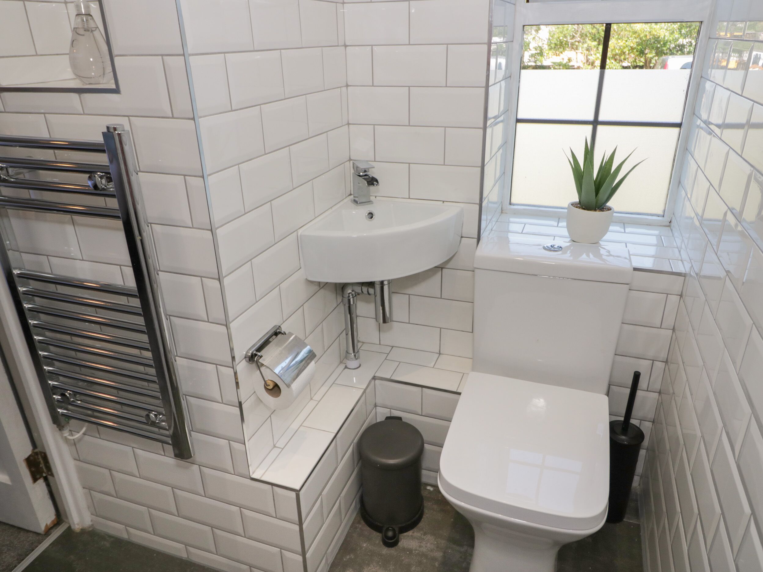 A bathroom with white tiled walls, a white sink, a toilet, a black trash bin, and a window with a potted plant.