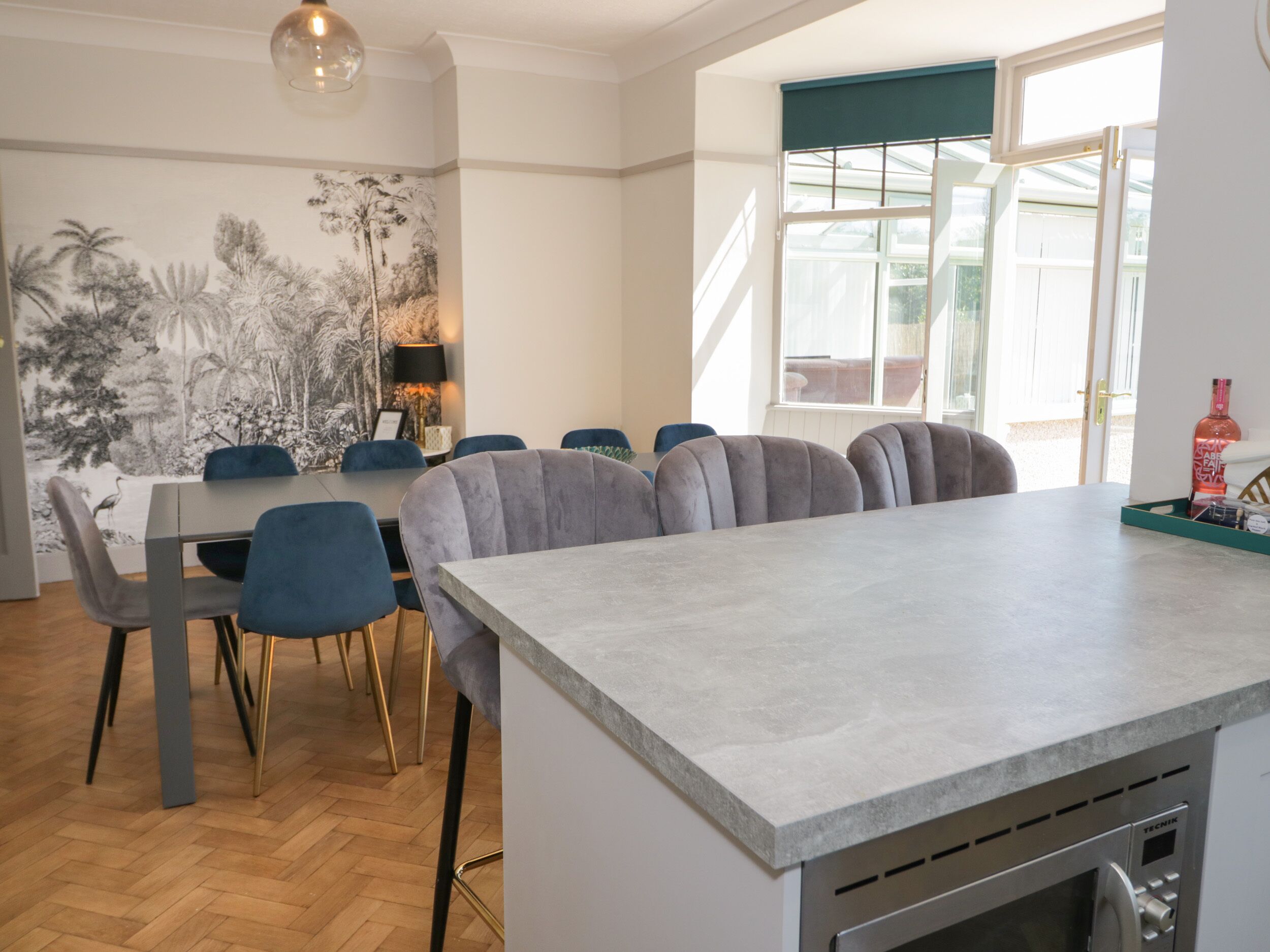 A modern kitchen with a central island, a dining area with blue chairs, and a large window with a view.