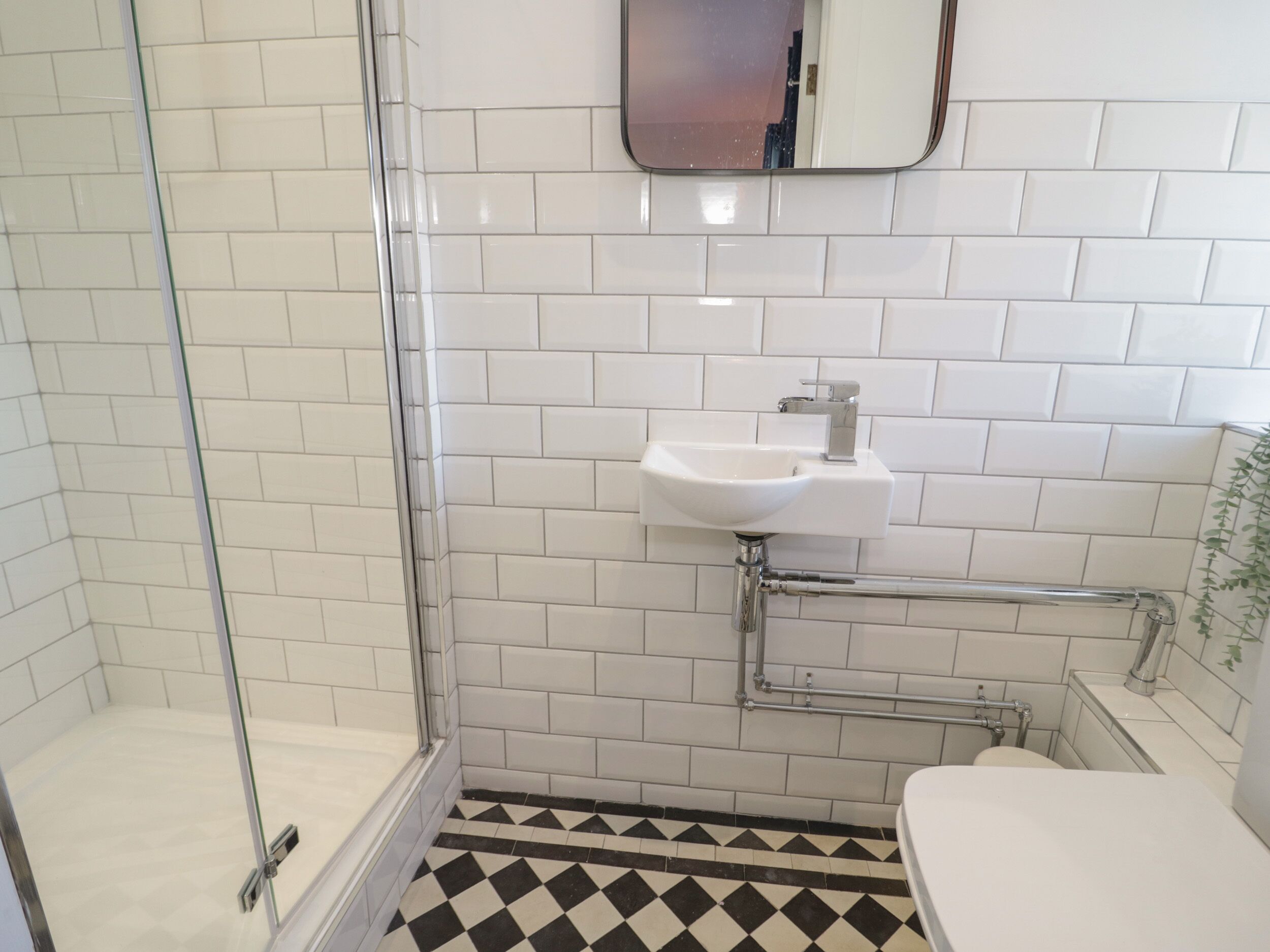 Une salle de bain avec des murs recouverts de carreaux blancs, un lavabo blanc, une cabine de douche en verre et un sol à carreaux noirs et blancs.