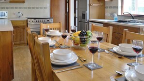 Un comedor con una mesa de madera dispuesta para cuatro personas, rodeada de sillas de madera. Hay un frutero sobre la mesa y copas de vino con vino tinto. Se ve la zona de la cocina al fondo, con armarios de madera y un refrigerador.