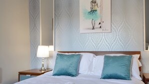 A hotel room with a bed, two pillows, a nightstand with a lamp, and a wall with a patterned wallpaper and a framed artwork. - Percy Park Apartment (TYNEMOUTH)
