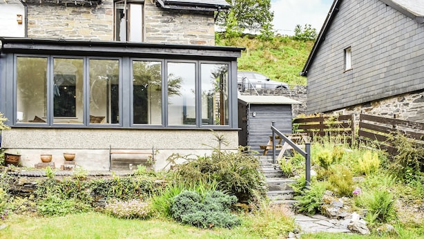 A stone house with a large glass window, a wooden deck, and a stone pathway leading to a door. - 2 bedroom accommodation in Dolwyddelan (Dolwyddelan)