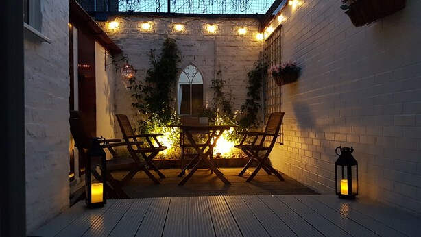 Patio esterno con tavolo e sedie in legno, circondato da pareti in mattoni bianchi e luci decorative.