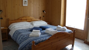 En træseng med hvidt og blåt sengetøj, et billede i ramme på væggen og et vindue med udsigt.