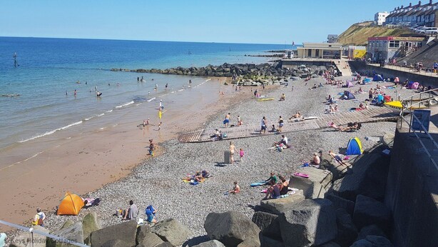 Une plage où des gens se prélassent au soleil et se baignent, un rivage rocheux et des bâtiments en arrière-plan.