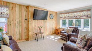 A living room with a wooden wall, a flat-screen TV, a clock, a window with a floral valance, and a brown leather sofa.