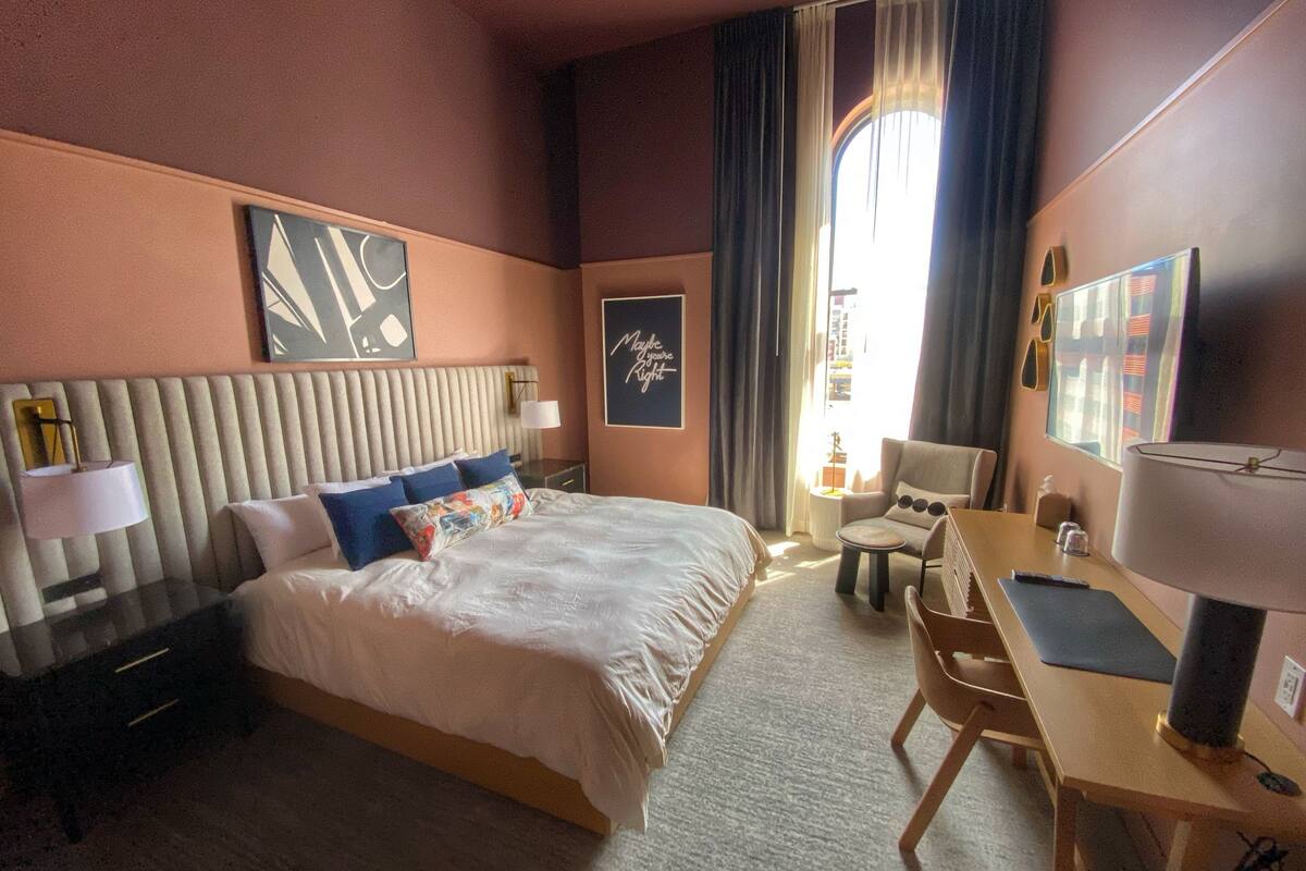 Une chambre d’hôtel avec un grand lit, un bureau, une chaise, une table de chevet et une fenêtre avec des rideaux.