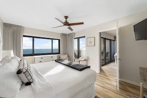 A hotel room with a large bed, a ceiling fan, a TV, and a balcony with a view of the sea.