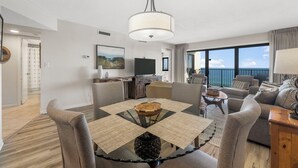 A modern living room with a glass-top dining table, grey sofas, and a large window offering a view of the ocean. - Emerald Towers 902 (Destin)