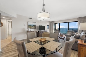 A modern living room with a glass-top dining table, grey sofas, a flat-screen TV, and a balcony with a view.