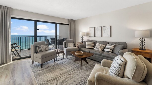 A modern living room with a sofa, armchairs, a coffee table, and a view of the ocean through large windows. - Emerald Towers 902 (Destin)
