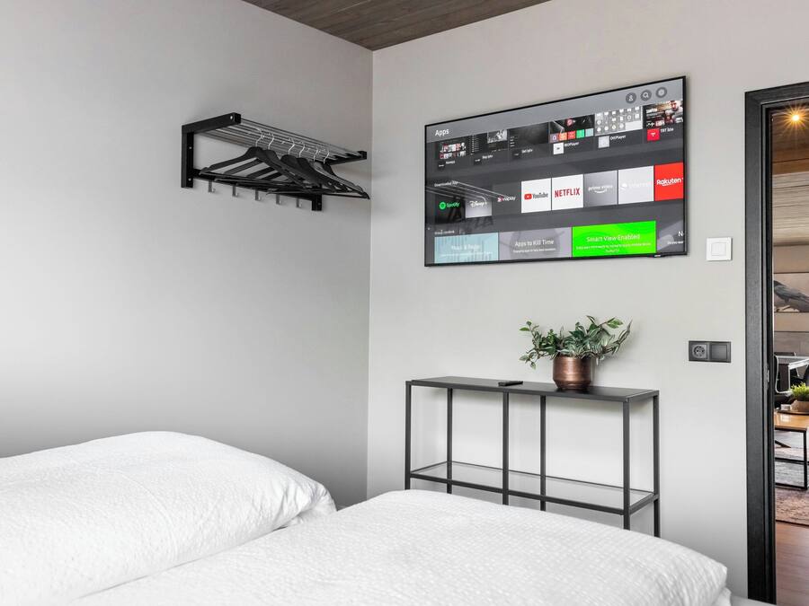 A modern hotel room with a bed, a wall-mounted TV displaying various streaming apps, a small table with a plant, and a coat rack.