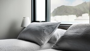 A bed with white bedding and pillows, a bedside lamp, and a window with a mountain view. - Pier Apartments (Vestmannaeyjar)