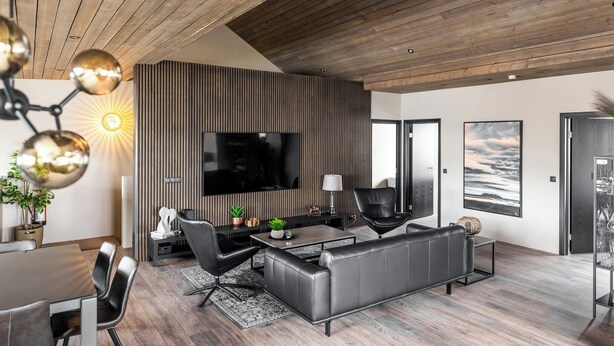 A modern living room with a large flat-screen TV, leather sofas, a dining area with a table and chairs, and a wooden ceiling. - Pier Apartments (Vestmannaeyjar)