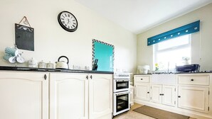 A kitchen with white cabinets, a black clock, a blue window blind, and a kettle on the counter. - Fourpenny Studio - UK34745 (Skelbo, near Dornoch)