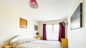 A bedroom with a bed, wooden nightstands, a bedside table, a red lamp, a window with curtains, and a wall-mounted TV. - Fourpenny Studio - UK34745 (Skelbo, near Dornoch)