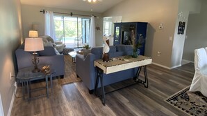 Uma sala de estar ampla, com um sofá azul, uma mesa de centro de vidro e um ventilador de teto.