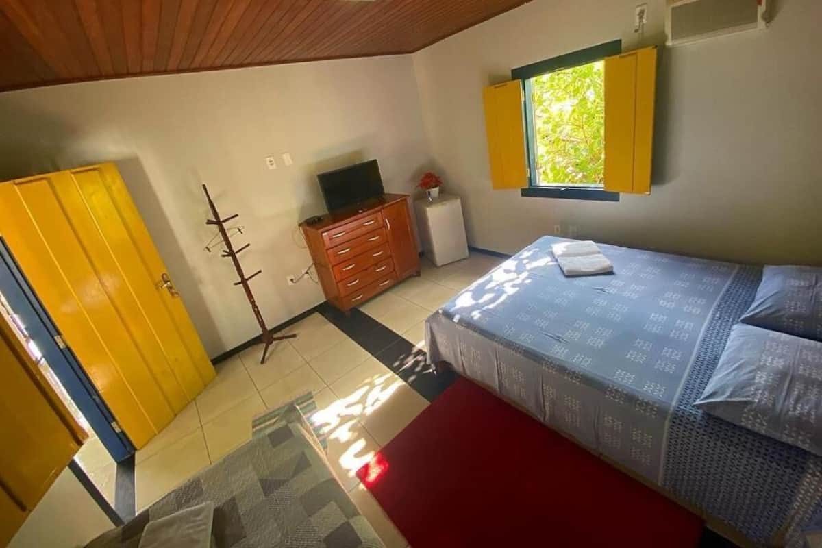 Quarto com cama, penteadeira, televisão e janela com persianas amarelas.