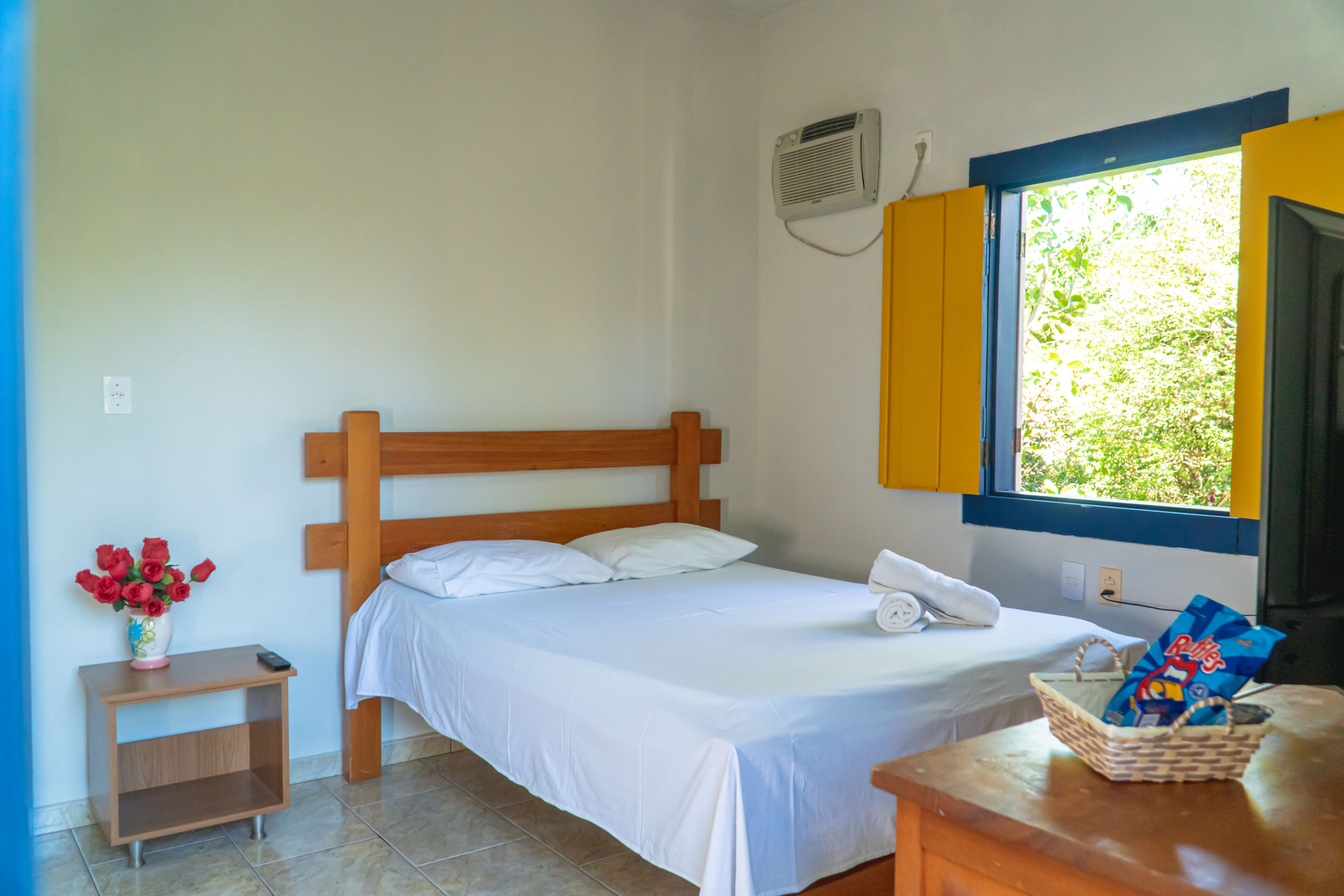 A neatly made bed with white linens, a bedside table with a vase of red roses, and a window with a view of greenery.